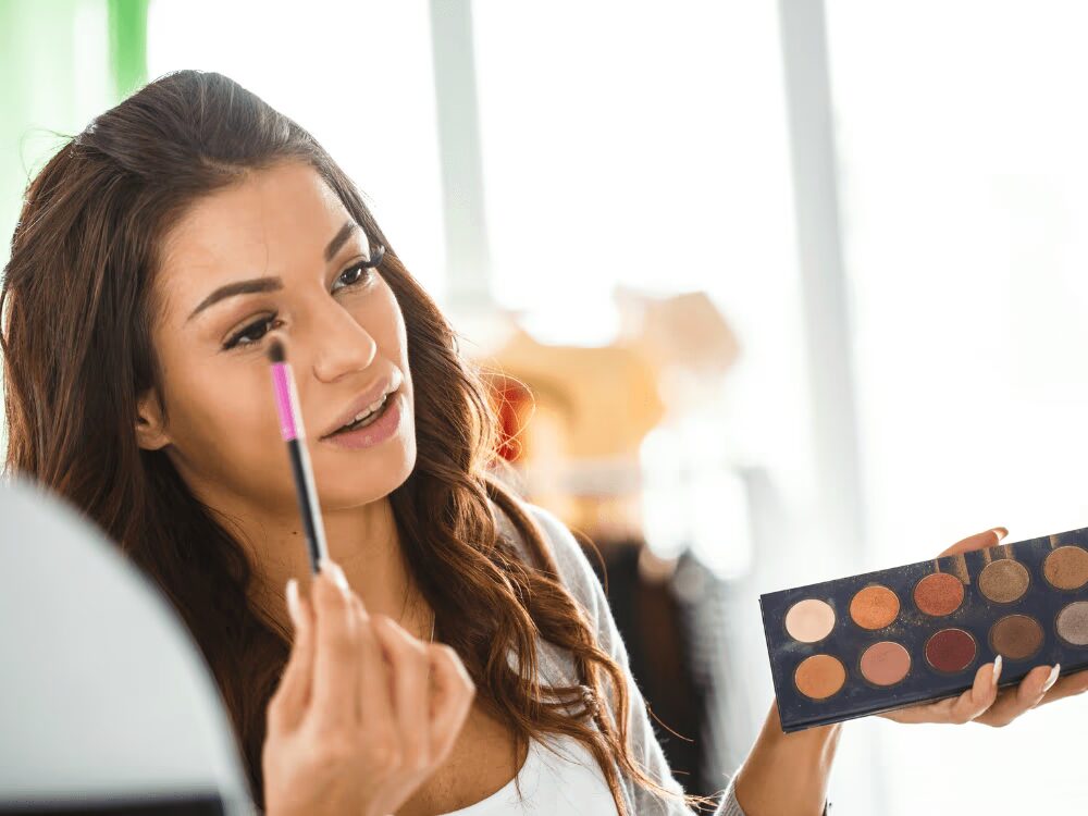 Mulher morena com cabelo liso, aplicando sombra nos olhos com pincel, em frente a um espelho pequeno, com paleta de maquiagem ao lado, em um ambiente com luz natural.