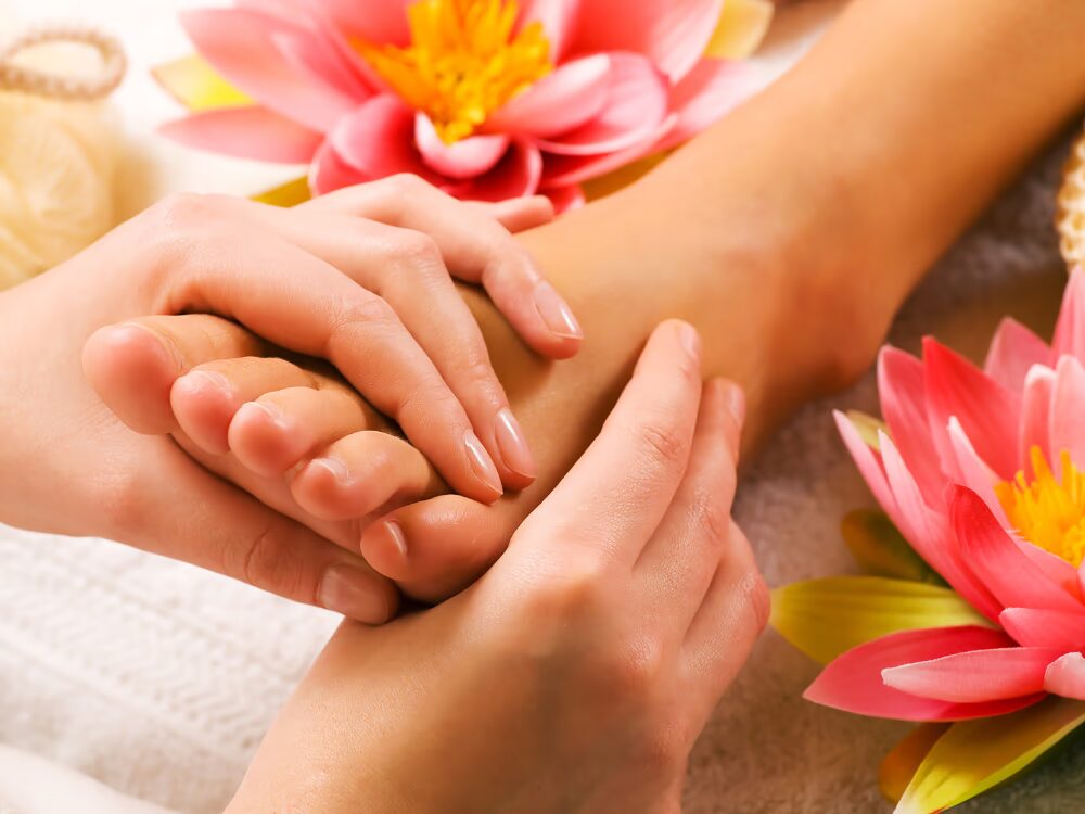 Massagem em um pé, com duas mãos aplicando pressão nos dedos e na planta. O pé está apoiado sobre uma toalha branca, cercado por flores de lótus rosa e amarelas, criando uma atmosfera relaxante típica de um spa. Ao fundo, há uma esponja natural, reforçando o ambiente de cuidados pessoais.