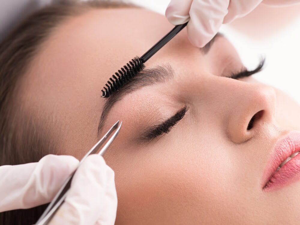 Close-up de um rosto feminino com olhos fechados, recebendo cuidados nas sobrancelhas por mãos com luvas brancas, usando uma pinça e um aplicador de máscara, em um ambiente com fundo claro.