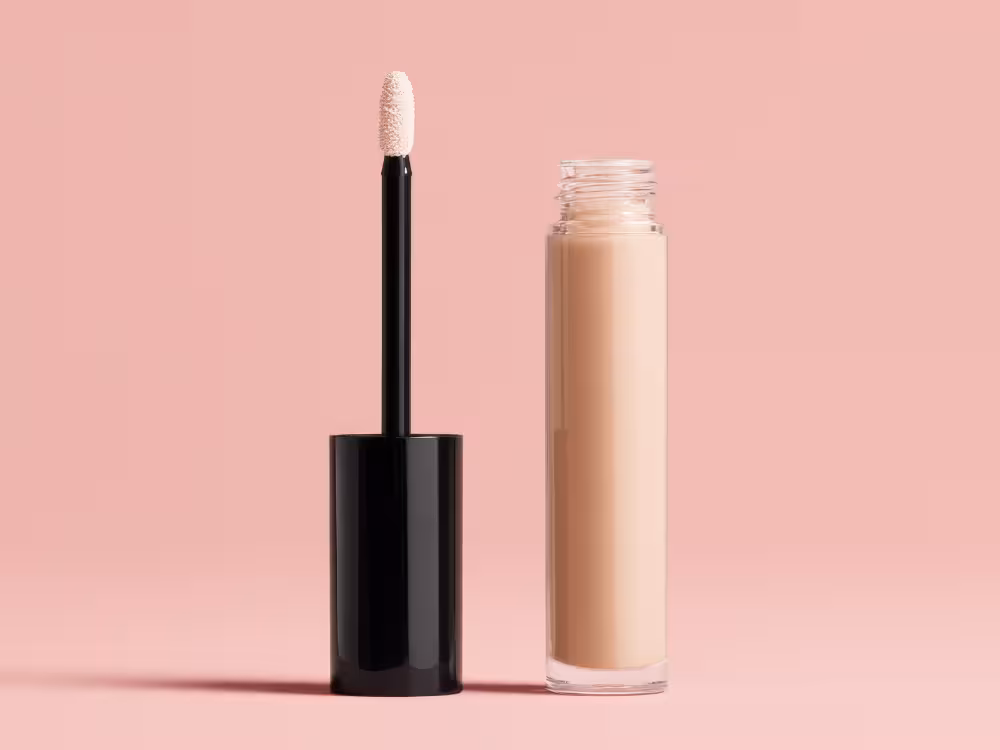 Corretivo líquido nude com aplicador esponja branca e embalagem preta transparente, aberto sobre fundo rosa pastel.