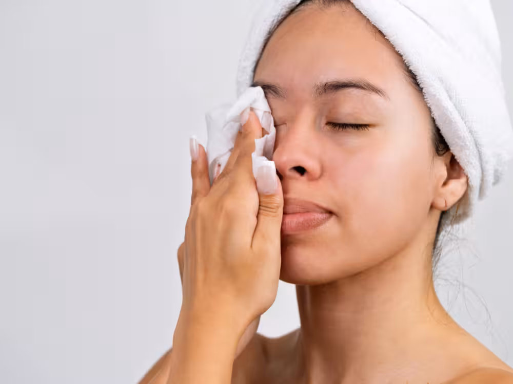 Mulher jovem de pele morena clara, com toalha branca enrolada na cabeça como turbante, pressionando delicadamente um lenço de papel branco sobre a região dos olhos fechados, provavelmente removendo maquiagem ou limpando o rosto após o banho. Expressão relaxada e serena, close-up do rosto com fundo branco neutro e iluminação suave.