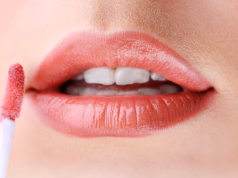 Close-up de aplicação de batom rosa brilhante com aplicador felpudo branco nos lábios, acabamento glossy e molhado, pele glowy natural.