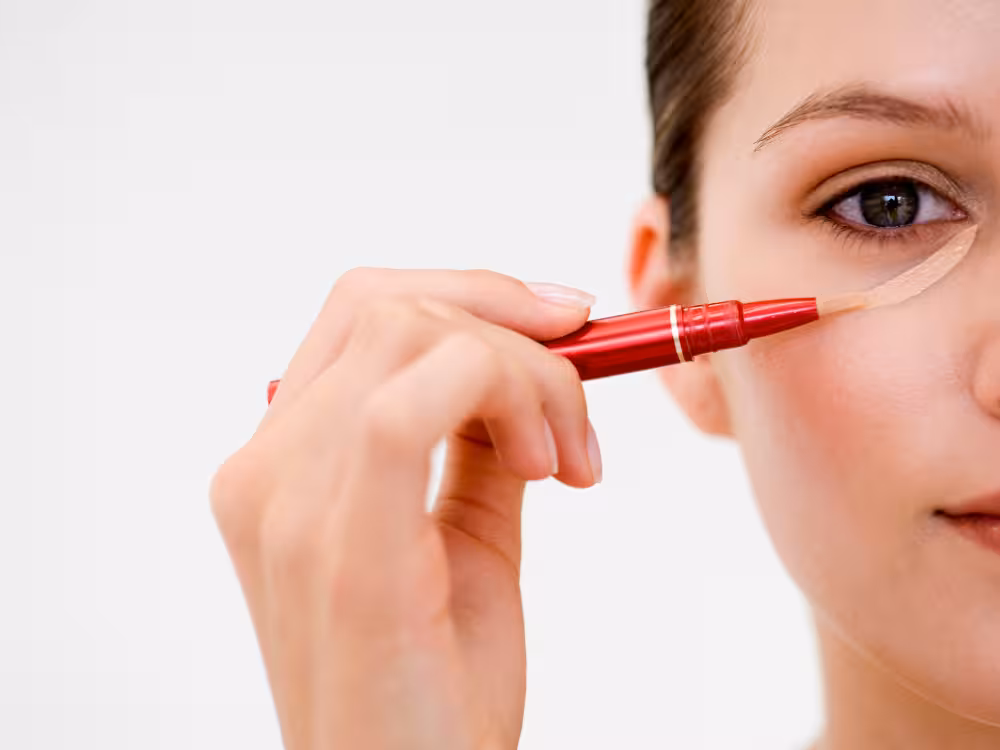 Mulher aplicando corretivo bege com bastão vermelho sob os olhos para cobrir olheiras, close-up de maquiagem natural.