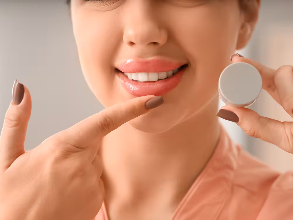 Mulher sorridente aplicando balm ou hidratante labial com o dedo no lábio inferior, segurando potinho cinza claro, maquiagem natural glowy e unhas marrons.