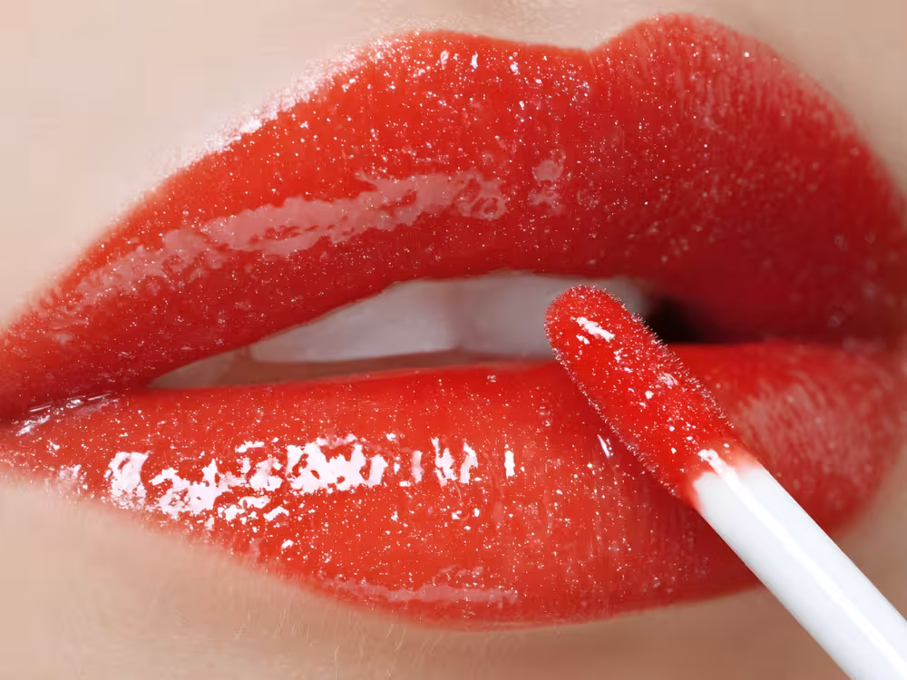Close-up de aplicação de batom vermelho brilhante com aplicador felpudo branco nos lábios, acabamento glossy intenso e molhado, fundo vermelho.