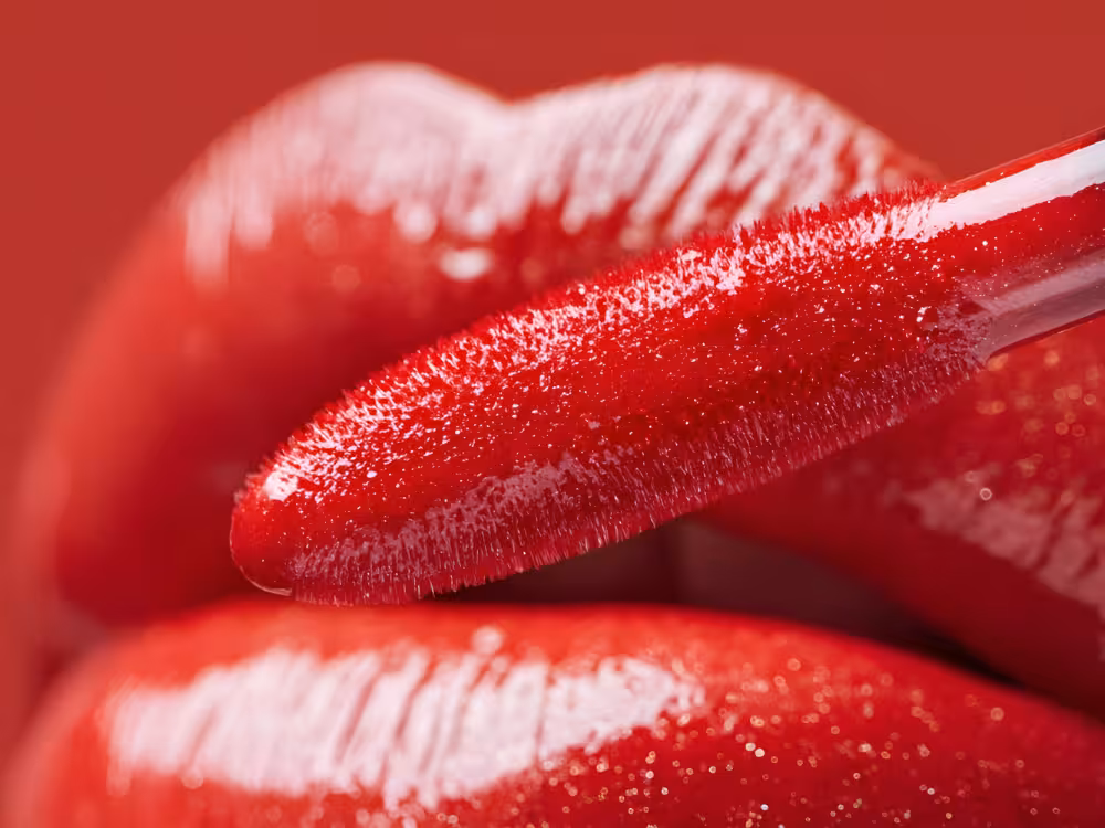 Close-up de aplicação de batom líquido vermelho brilhante com aplicador felpudo nos lábios, acabamento glossy e intenso, fundo vermelho.