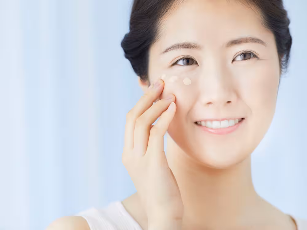 Mulher asiática aplicando corretivo bege em pontos sob o olho com o dedo, maquiagem natural glowy, sorriso leve, fundo azul claro.