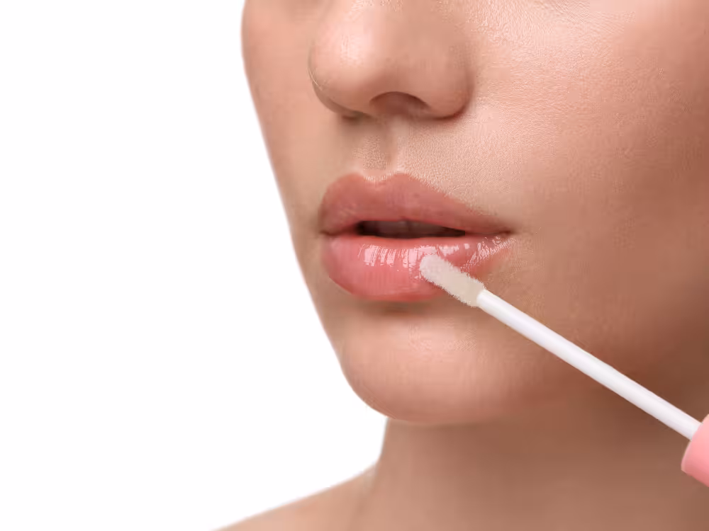 Mulher aplicando gloss nude-rosado com aplicador felpudo branco nos lábios, close-up de boca com acabamento brilhante e molhado.