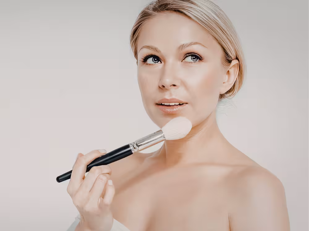 Mulher loira aplicando blush ou pó com pincel na maçã do rosto, close-up de rosto glowy natural com olhos azuis e lábios nude, fundo bege.