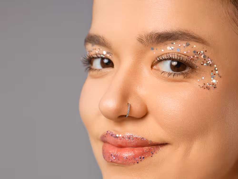 Mulher com maquiagem de glitter colorido em arco nos olhos e nos lábios rosa brilhante, piercing de septo, close-up de rosto com expressão serena.