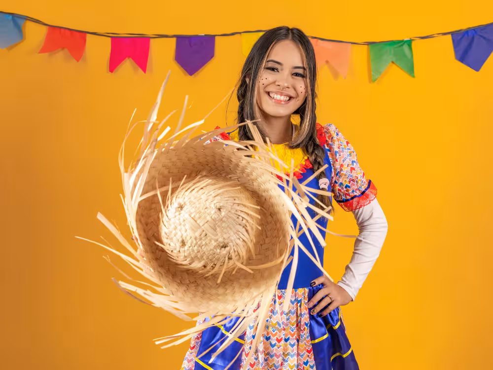 Mulher sorridente fantasiada de quadrilha junina com vestido colorido, chapéu de palha grande e sardas pintadas no rosto, bandeirinhas coloridas ao fundo amarelo.