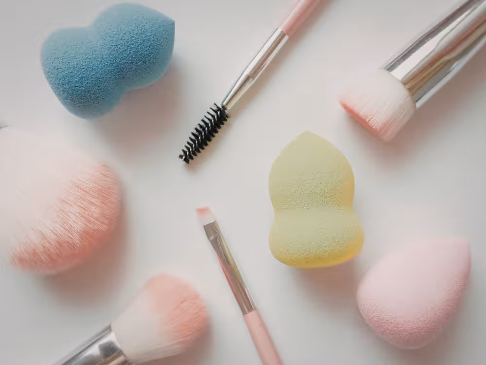 Flat lay de esponjas de maquiagem coloridas (coração azul, beauty blender verde e rosa), pincel rosa e escovinha de rímel preta, fundo bege claro.