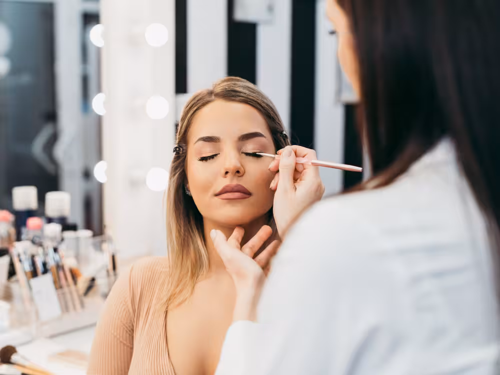Maquiadora aplicando sombra ou delineador nos olhos de mulher sentada em penteadeira iluminada, olhos fechados, close-up de rosto com make em andamento.
