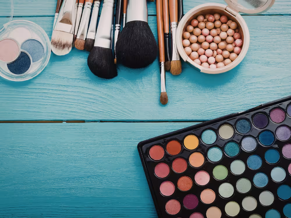 Flat lay de maquiagem sobre fundo turquesa: paleta de sombras colorida, pincéis variados, esponja de bolinhas e paleta redonda de sombras azuis e rosas.