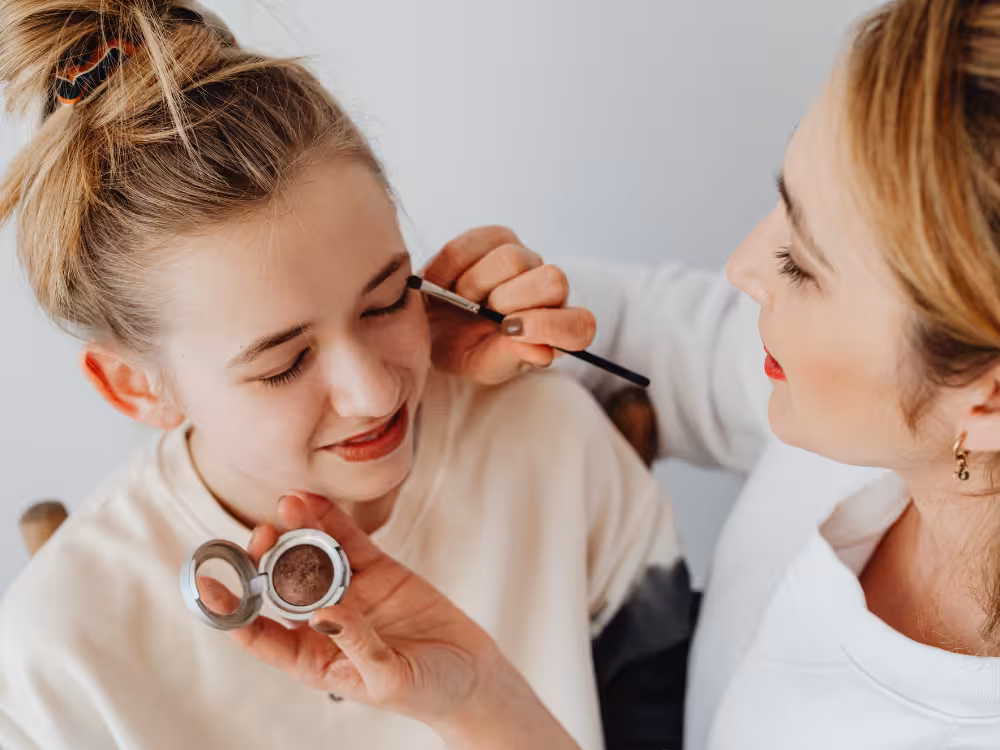 Mulher aplicando sombra ou produto de sobrancelha com pincel na adolescente loira, que segura compacto marrom e sorri com olhos fechados, fundo claro.