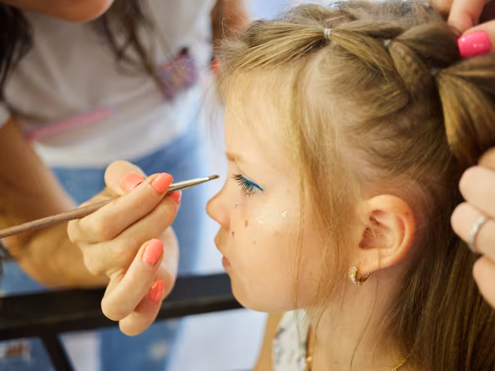 Menina com tranças recebendo maquiagem facial: delineado azul e glitter nas bochechas, pincel sendo aplicado no olho por adulta, close-up de rosto.