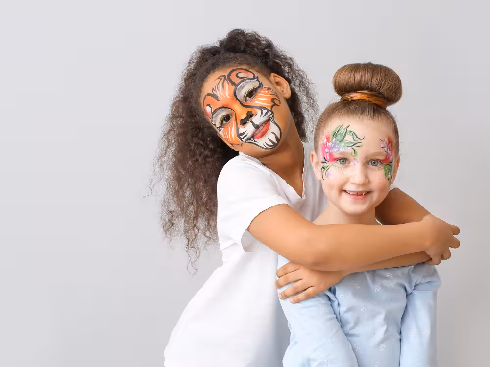 Duas meninas abraçadas com face painting: uma como tigre laranja e preto, outra com flores coloridas no rosto, sorrisos largos, fundo cinza claro.