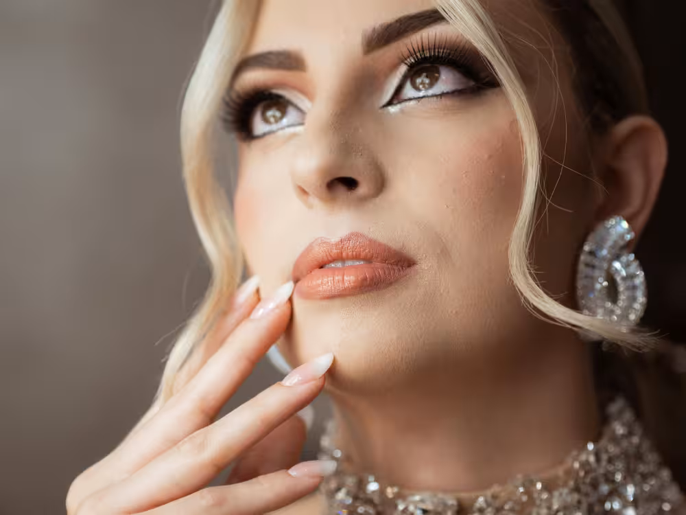 Mulher loira com maquiagem glam: sombra esfumada dourada e glitter, delineado preto, batom nude brilhante, brincos e colar cravejados, mão no queixo, close-up de rosto glowy.