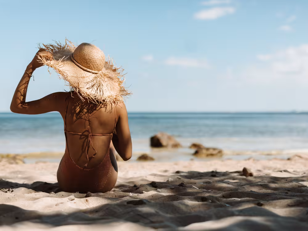 Mulher de costas sentada na areia da praia, de maiô e chapéu de palha grande, admirando o mar. Imagem ilustra um dia de sol e a importância de maquiagem resistente à água e com proteção solar para o verão.