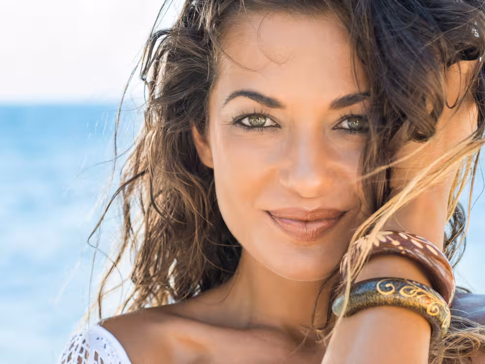 Close-up de mulher com olhos verdes e cabelos cacheados na praia, com maquiagem natural e bronzeada. Ela sorri para a câmera com o oceano ao fundo, ilustrando um look de verão leve e fresco.