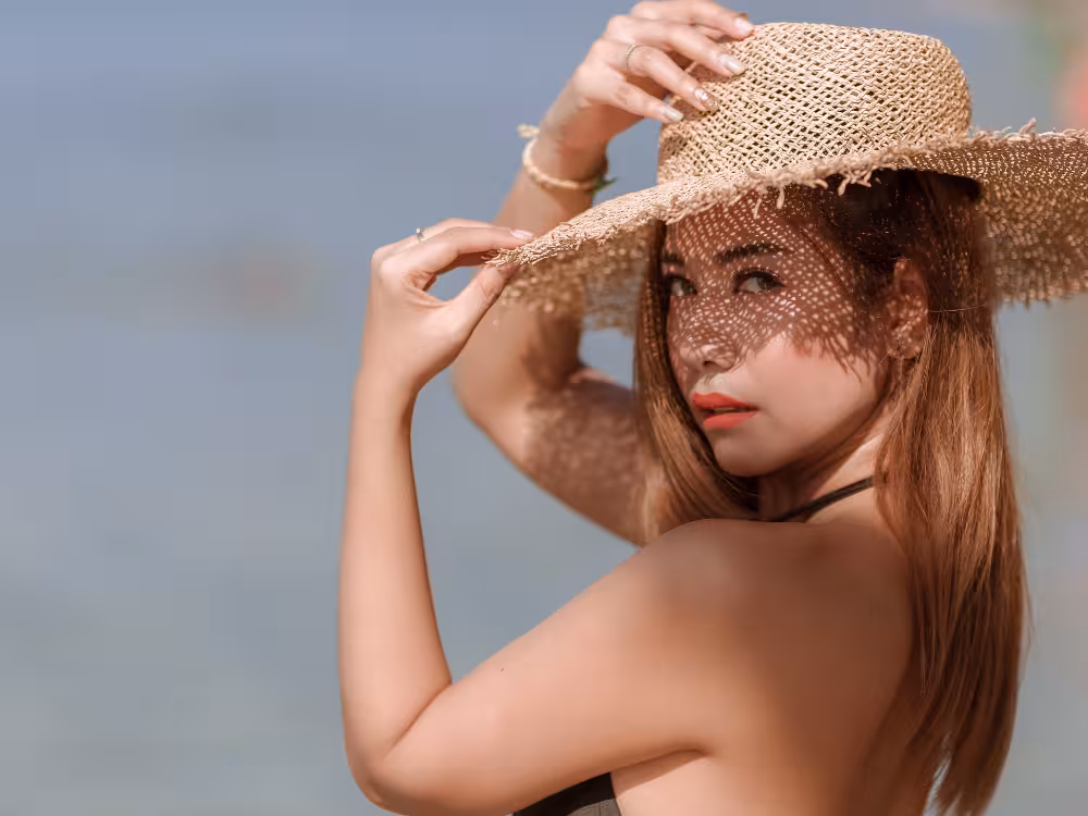 Close de mulher na praia segurando um chapéu de palha de aba larga sobre a cabeça. Ela usa um batom coral vibrante e maquiagem de olhos leve, com sombras texturizadas projetadas pelo chapéu em seu rosto. O fundo é um mar azul desfocado em um dia ensolarado, ilustrando um look de maquiagem para o verão.