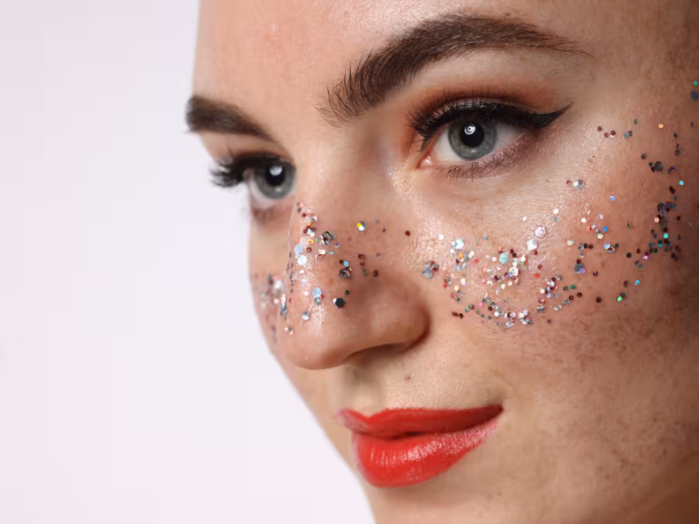 Mulher com glitter holográfico multicolorido aplicado no nariz e maçãs do rosto, sombra laranja brilhante, delineado gatinho e batom vermelho, close-up de make festiva.