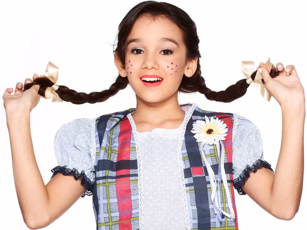 Menina em fantasia junina com colete xadrez colorido, tranças com laços bege, sardas de coração no rosto, flor no peito e sorriso animado, fundo branco.