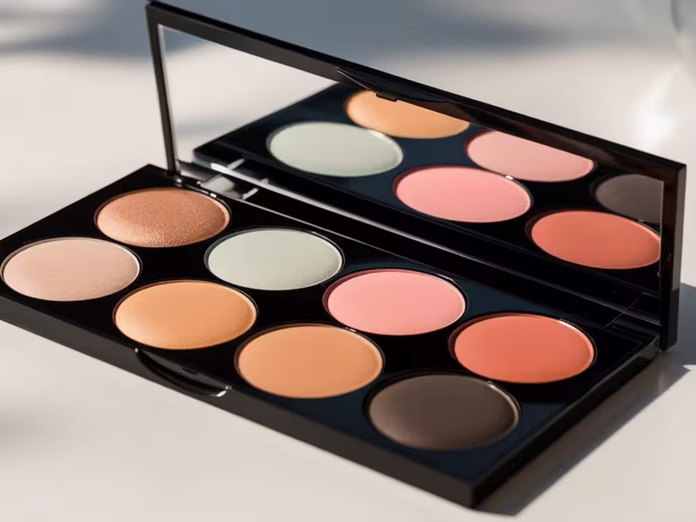 Paleta de maquiagem preta aberta com dez cores: iluminador dourado, blushes rosados e pêssego, corretivo verde e tom marrom para contorno, sobre fundo branco.