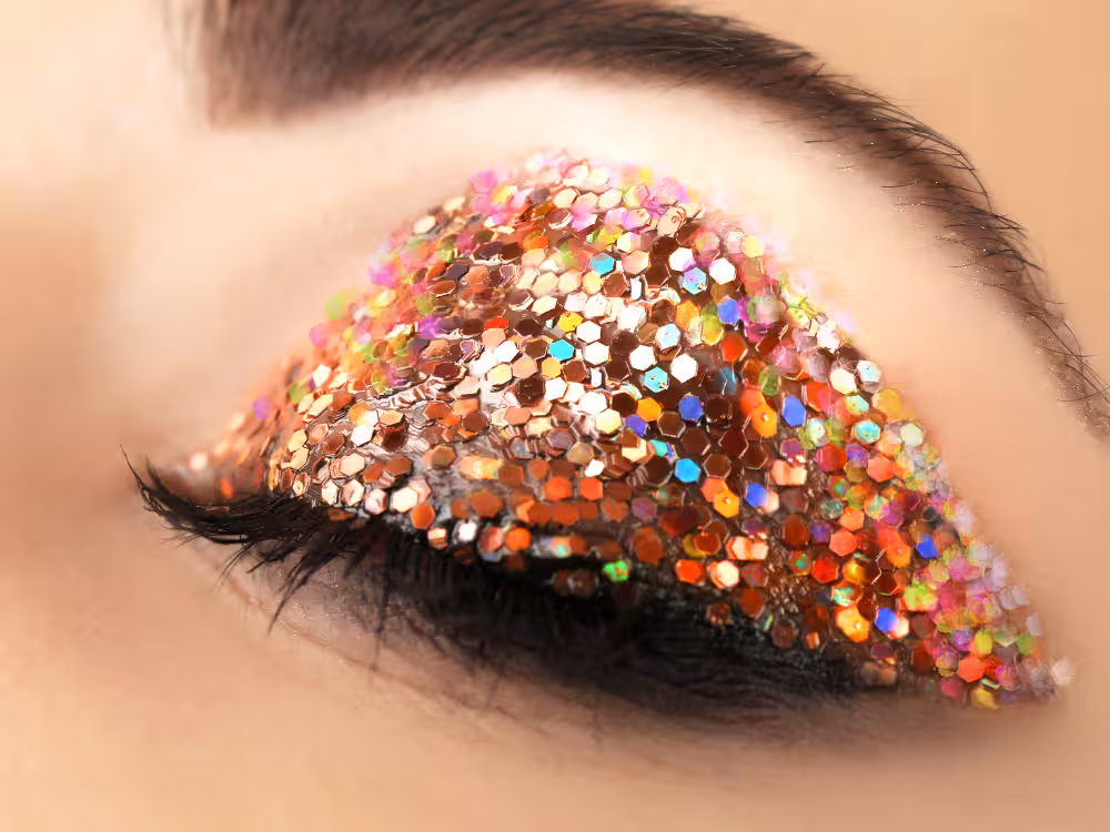 Close-up de olho com maquiagem de glitter holográfico multicolorido em formato de asa, tons cobre, rosa e arco-íris, cílios longos e sobrancelha marcada.