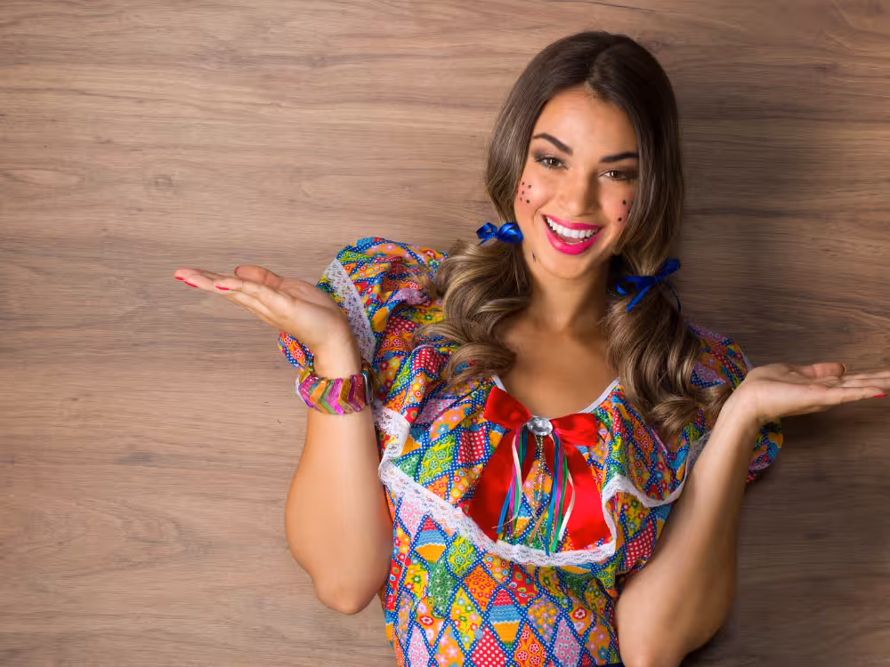 Mulher sorridente em traje junino colorido rodado, sardas de coração no rosto, laços azuis no cabelo, braços abertos em pose festiva, fundo de madeira clara.