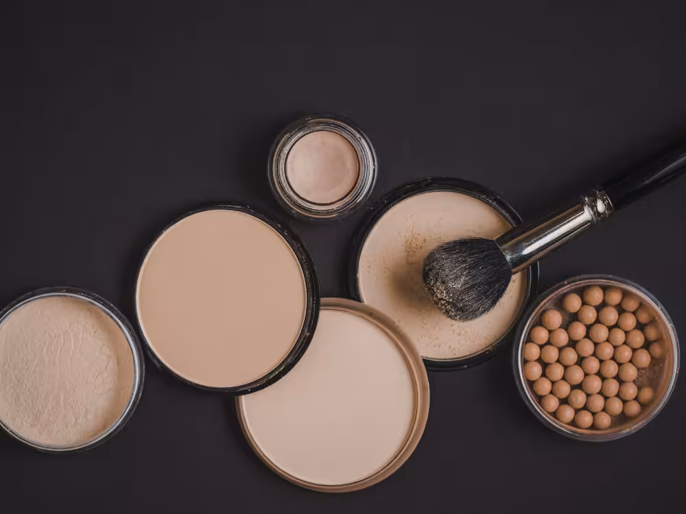 Visão superior de diversos tipos de pó facial: dois pós compactos, um pó translúcido solto, um potinho com pó iluminador e pó em pérolas bronzeadoras, com um pincel de maquiagem preto sobre a superfície escura.