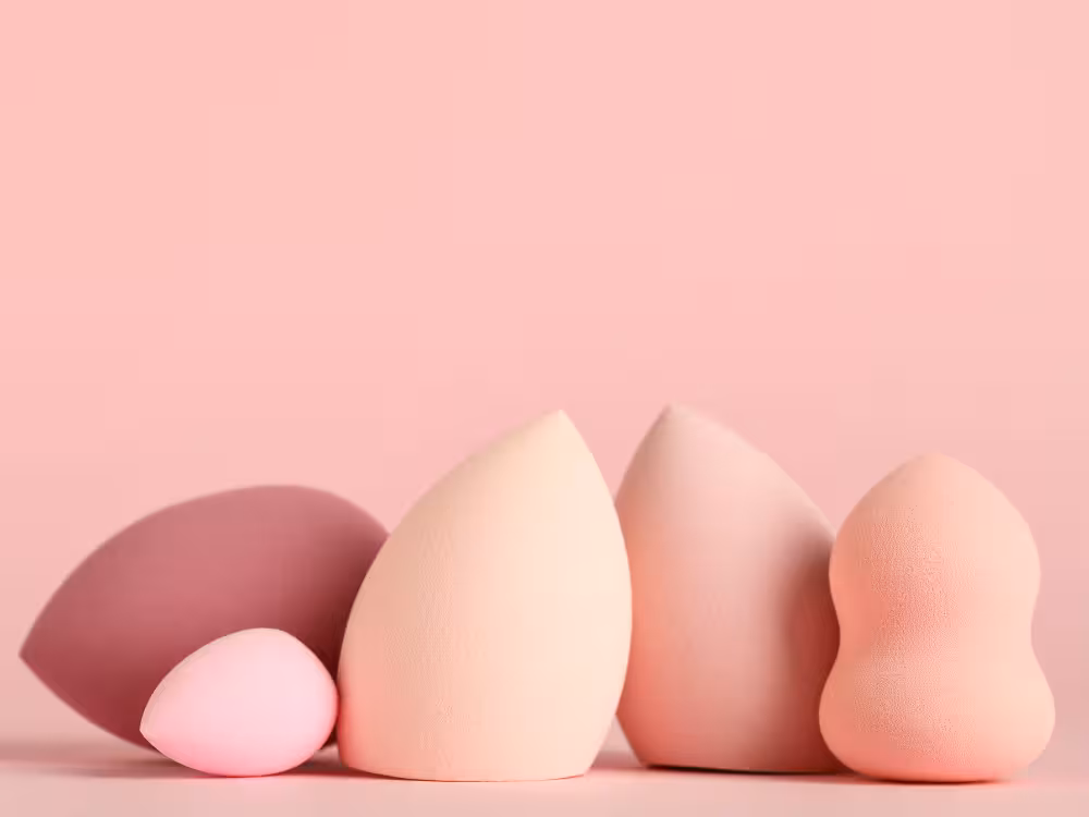 Flat lay de beauty blenders em tons de rosa pastel e bege claro, dispostas sobre fundo rosa suave, composição minimalista de esponjas de maquiagem.
