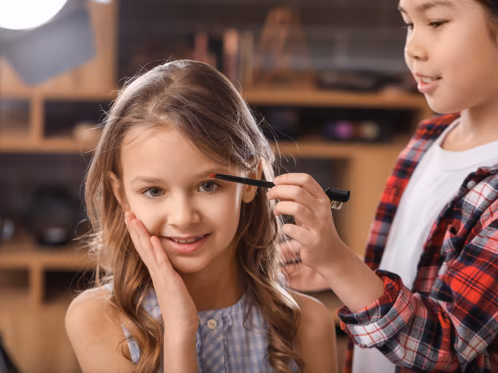 Menina sorridente recebendo maquiagem infantil: outra criança aplicando lápis vermelho na sobrancelha com pincel, ring light ao fundo, ambiente de brincadeira.
