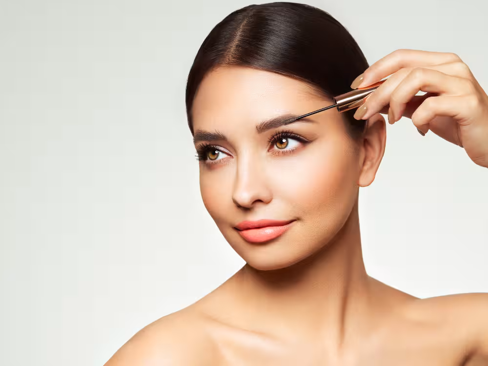 Mulher com pele glowy aplicando produto na sobrancelha com pincel fino dourado, maquiagem elegante com delineado marcado e batom nude, close-up de rosto.