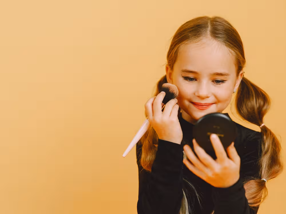 Menina sorridente aplicando blush ou pó com pincel rosa no rosto, olhando-se no espelhinho preto, tranças soltas, fundo laranja claro.
