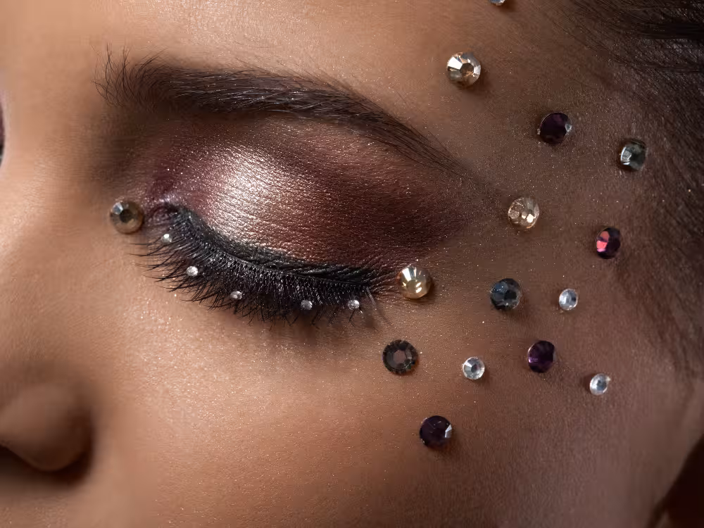 Close-up de olho fechado com maquiagem artística: sombra metálica rosê com esfumado vinho na pálpebra e cílios postiços volumosos. Strass em tons de dourado, prateado, roxo e grafite espalhados ao redor do olho e pela bochecha, criando efeito decorativo festivo. Pele morena escura.