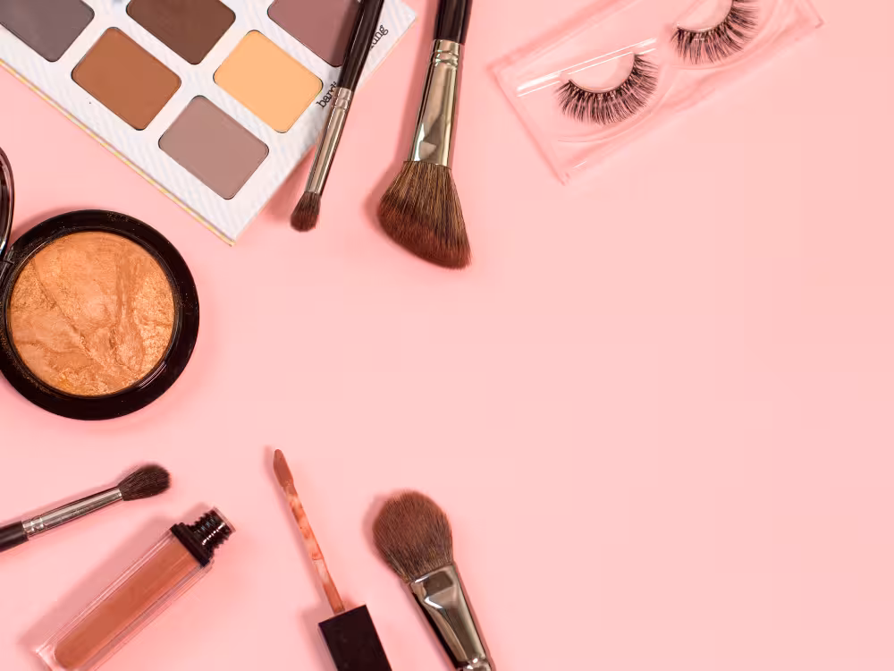 Uma vista superior de um kit de maquiagem para a praia essencial sobre um fundo rosa claro, incluindo uma paleta de sombras neutras, um bronzer compacto, um gloss labial nude, cílios postiços e vários pincéis. A imagem ilustra os produtos necessários para um visual natural e bronzeado de verão.