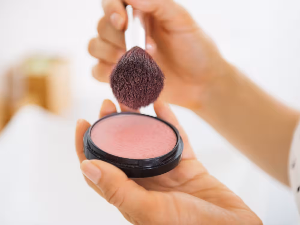 Close de mãos femininas segurando um estojo aberto de blush em pó rosado com relevo floral. Um pincel de maquiagem com cerdas fofas está pegando o produto, ilustrando como aplicar blush de forma leve para um visual natural de praia.