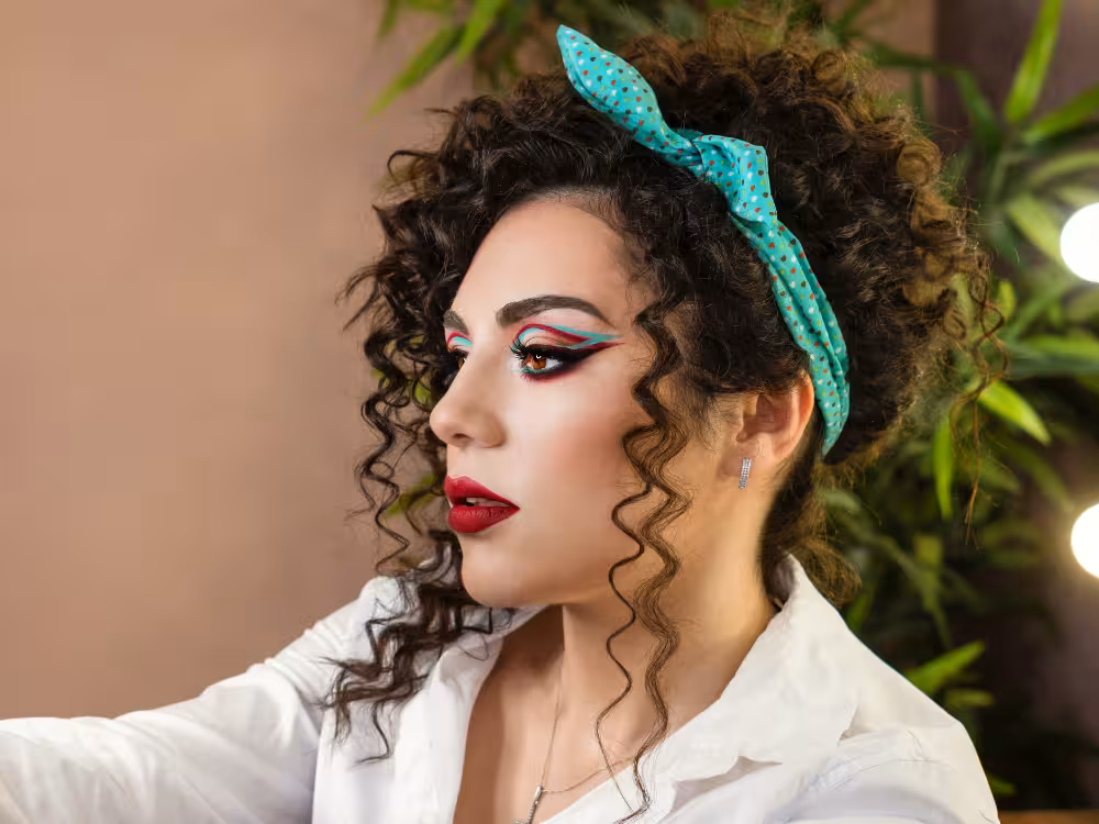 Mulher jovem de perfil com cabelos cacheados escuros presos por uma faixa de tecido verde-tiffany com poás. Maquiagem artística com delineado duplo em vermelho e turquesa estilo gráfico, cílios volumosos e batom vermelho escuro. Blusa branca, brinco prateado e colar fino. Fundo bege com folhagem desfocada.