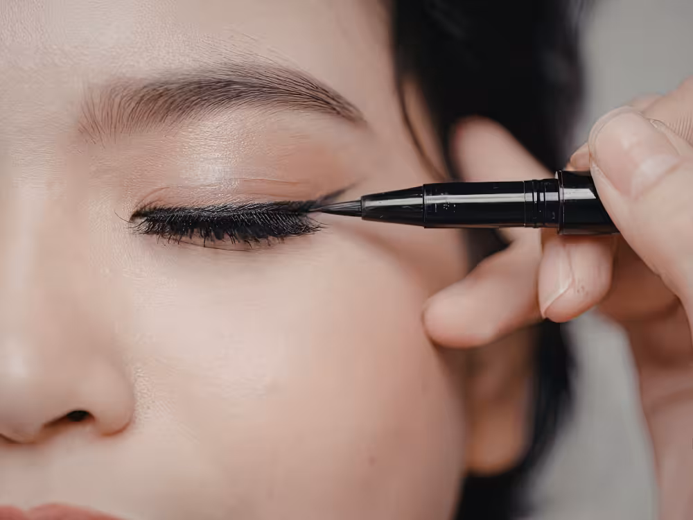 Close-up de mulher jovem de cabelos escuros e olho fechado, com delineador preto sendo aplicado na pálpebra superior com uma caneta delineadora de ponta fina. O traço forma um esfumado nas cílios, com fundo neutro e desfocado.