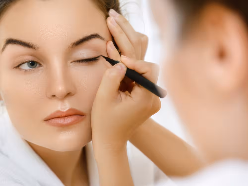 Maquiadora aplicando delineador líquido preto na pálpebra superior de uma mulher jovem de olhos claros e pele clara, vestindo roupão branco. A modelo mantém o olho semicerrado enquanto a profissional segura o lápis com precisão. Fundo claro e desfocado.