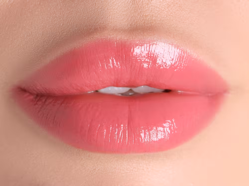 Close-up de lábios femininos com batom rosa médio e acabamento glossy brilhante, levemente entreabertos. Pele clara ao redor. Imagem focada nos lábios para destaque do produto.