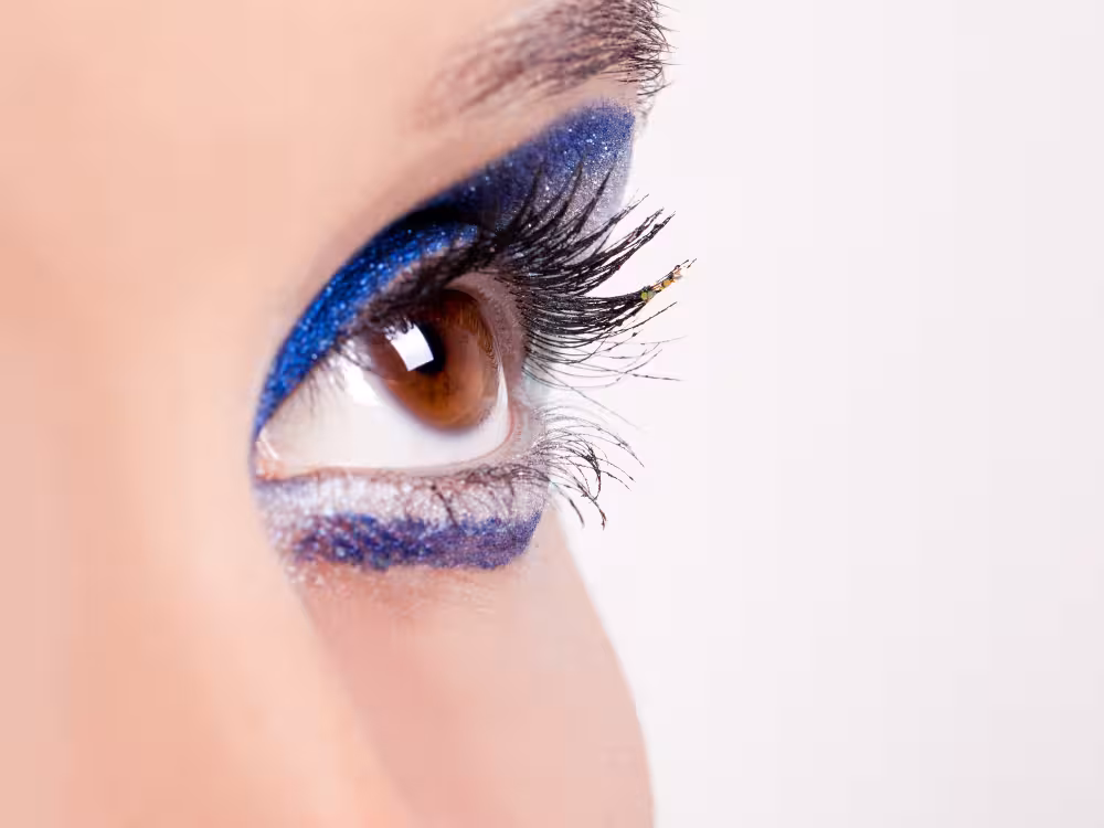 Close-up de olho aberto com íris castanha, maquiagem com sombra azul marinho com glitter na pálpebra superior e delineado azul com glitter prateado na linha d'água inferior. Cílios postiços alongados e volumosos. Pele clara e fundo branco desfocado.