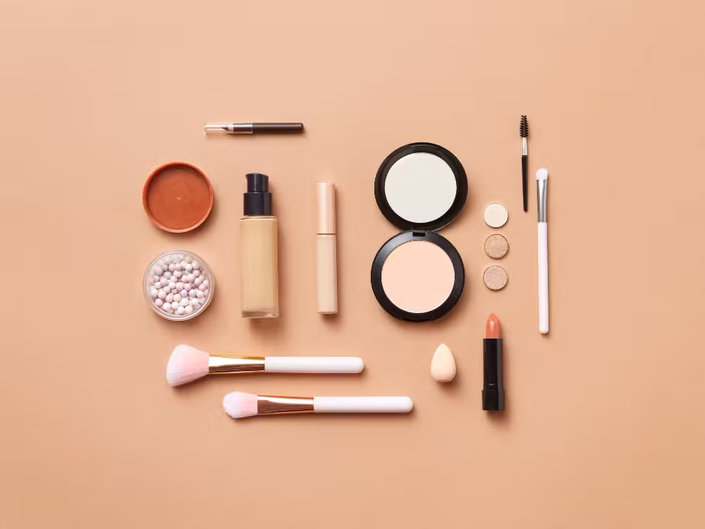 Visão superior de um kit essencial de maquiagem em tons neutros sobre fundo bege, contendo base líquida, corretivo, pó compacto, batom nude, pérolas iluminadoras e pincéis fofos.