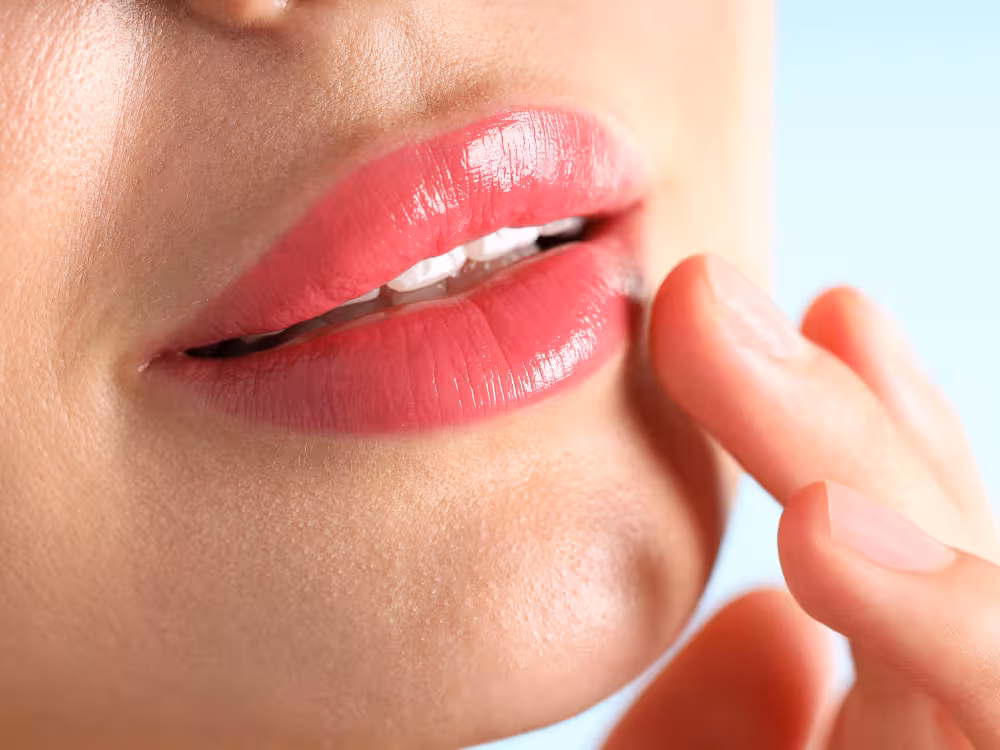 Close-up de lábios femininos entreabertos com batom rosa intenso e acabamento glossy brilhante, com dedos tocando o canto da boca. Pele clara e fundo azul claro desfocado.