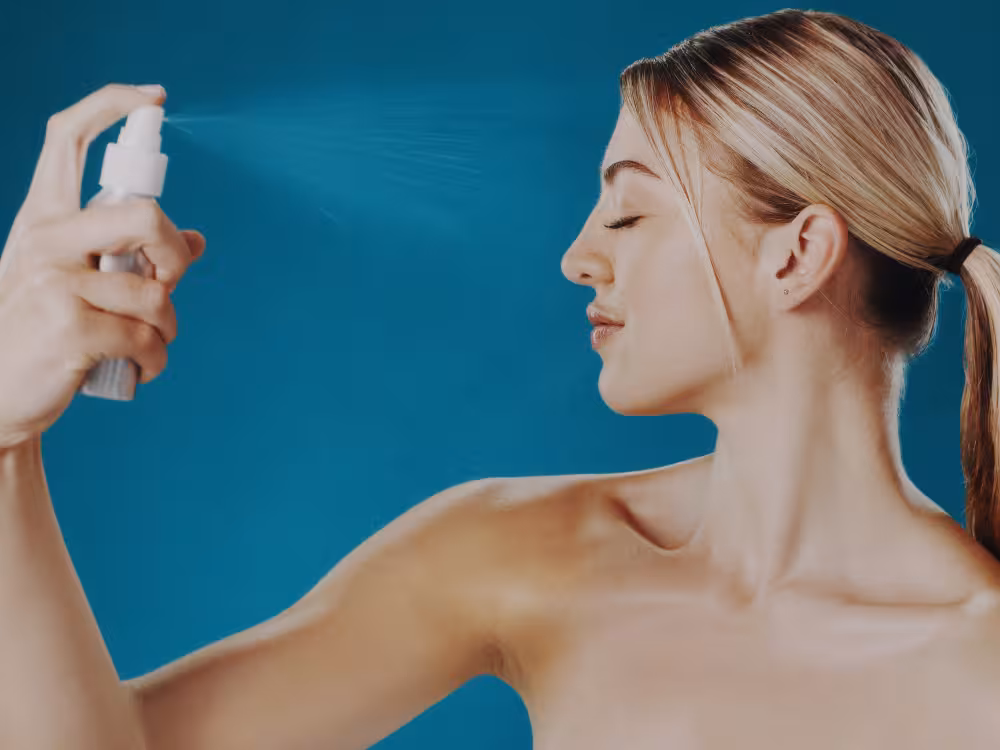 Close-up do rosto de uma mulher com maquiagem natural de praia, aplicando um batom ou lip balm nude rosado com acabamento hidratante nos lábios. A imagem ilustra o passo final para um look de verão leve e fresco.
