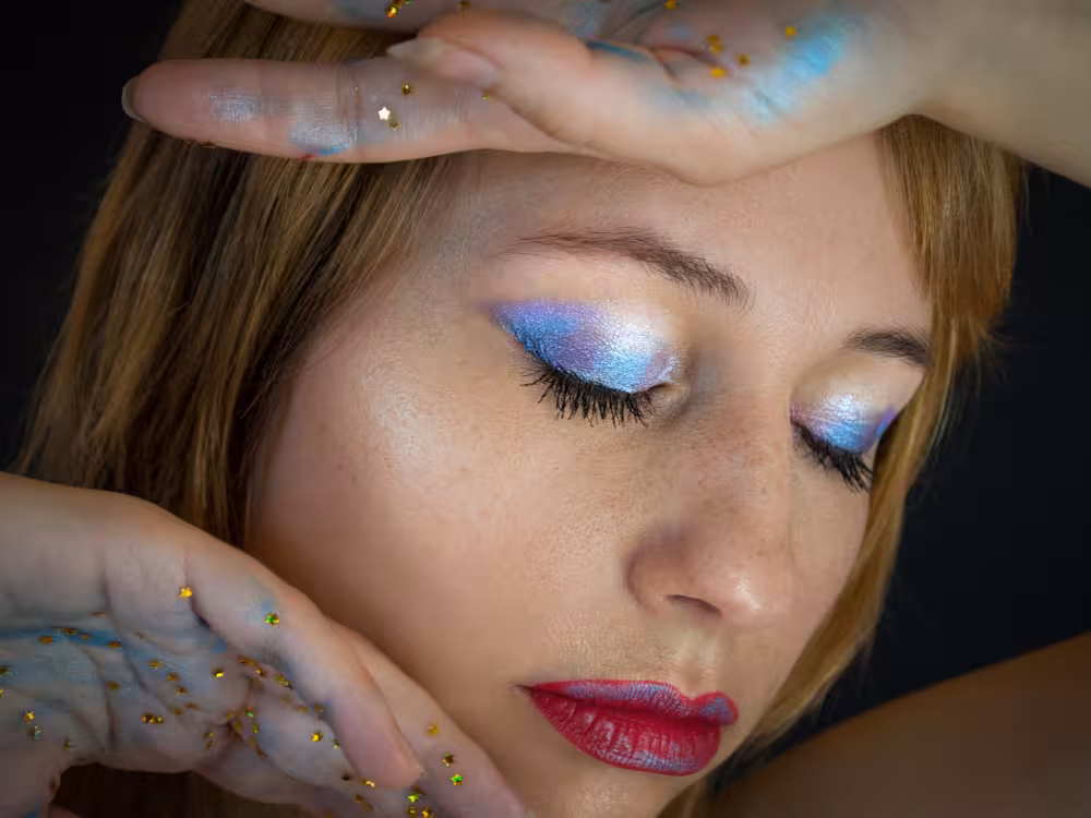 Mulher jovem de cabelos loiros com olhos fechados e maquiagem artística: sombra azul holográfica metálica nas pálpebras e batom vermelho intenso nos lábios. As mãos emolduram o rosto, com glitter em formato de estrelinhas douradas espalhadas pela pele. Fundo escuro com iluminação dramática.