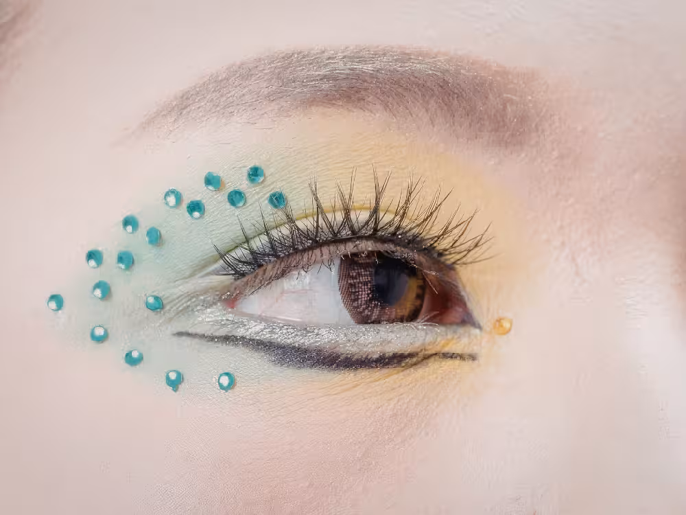 Close-up artístico de olho aberto com maquiagem criativa: sombra verde-menta e amarela na pálpebra, delineador prateado na linha d'água e diversos strass turquesa espalhados ao redor do olho em formato decorativo. Um strass dourado aparece no canto externo. Íris castanha com cílios alongados.