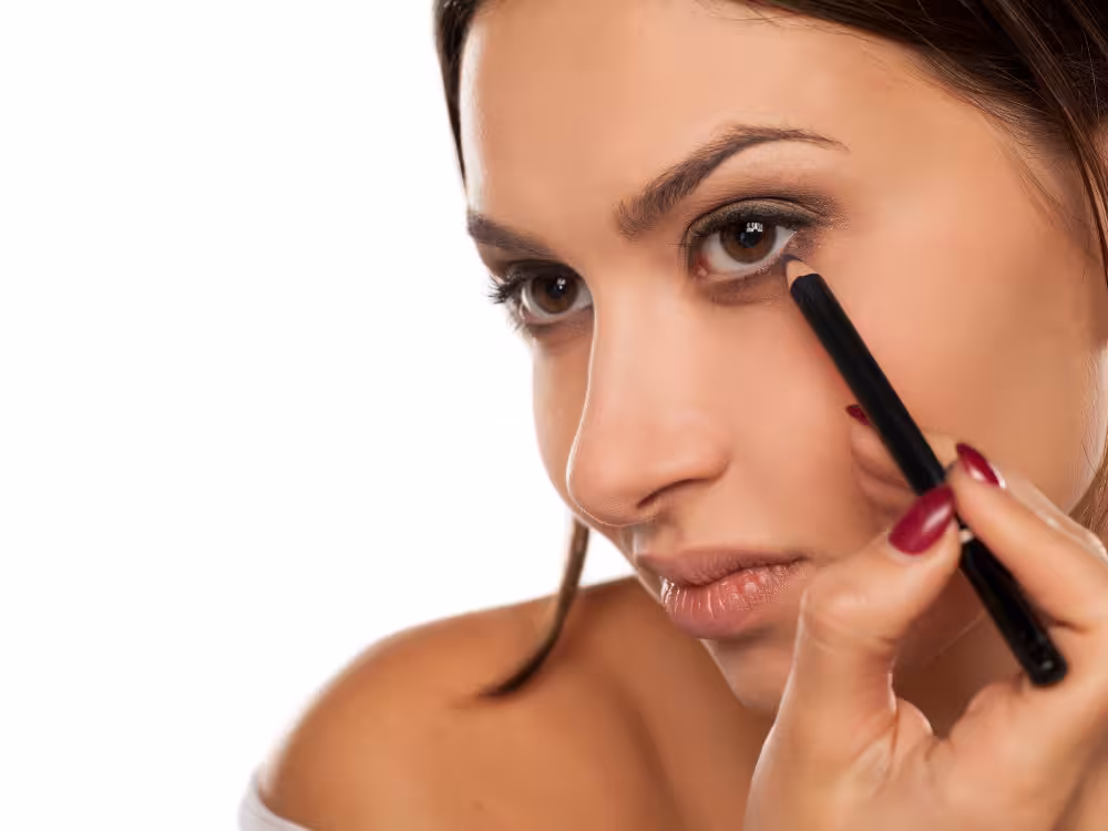 Mulher aplicando um lápis de olho preto na linha d'água inferior. Ela está levemente inclinada para o lado, olhando em direção à câmera, enquanto segura o lápis com precisão. Suas unhas estão pintadas de um tom vinho escuro e o fundo da imagem é branco e limpo.