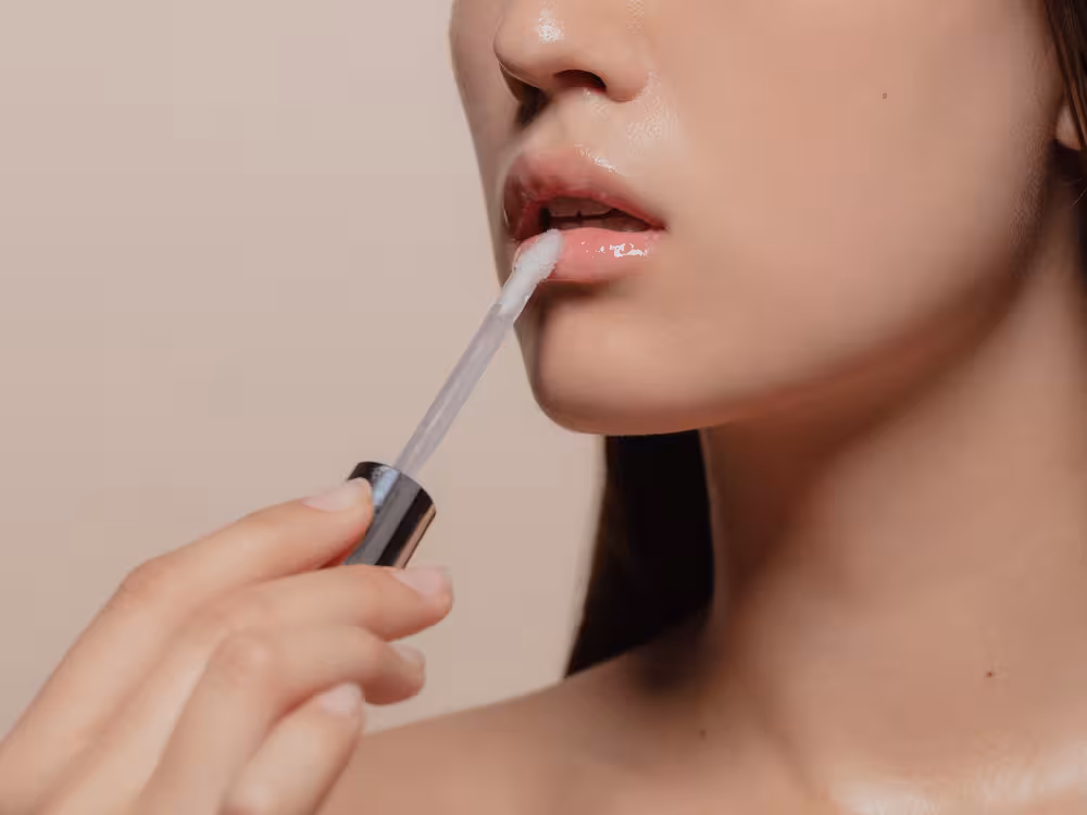 Close-up do rosto de perfil de mulher jovem de cabelos escuros aplicando gloss labial transparente nos lábios com o aplicador. Pele clara e luminosa. Fundo bege neutro.